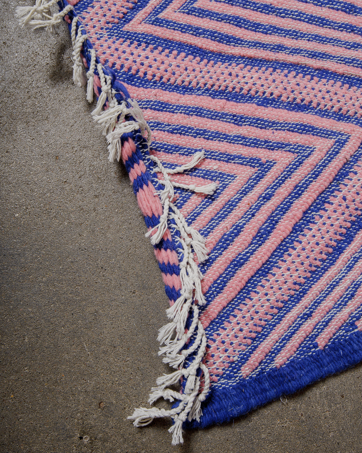 Close-up of a pink and blue woven rug with curled up, fringed edge on a concrete floor.